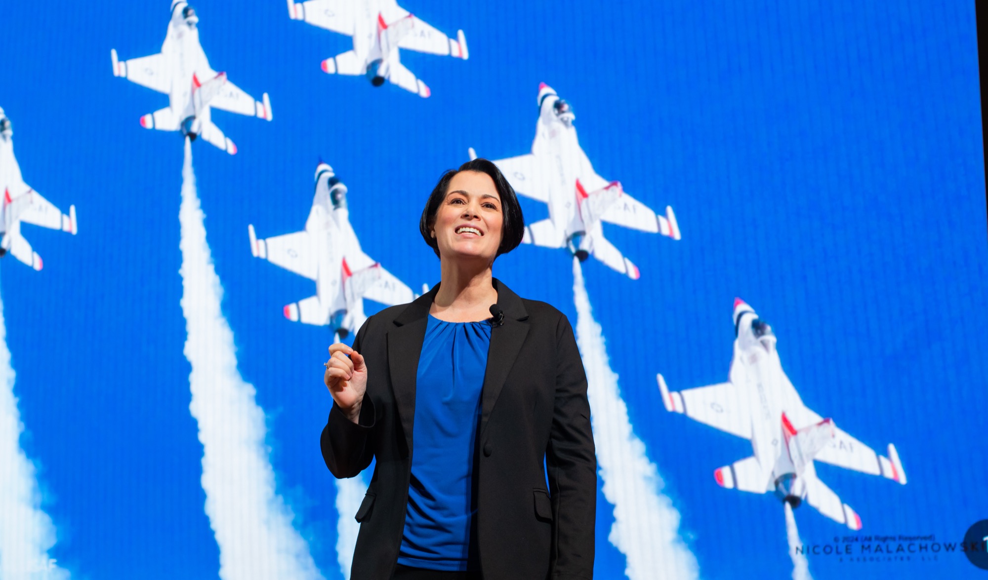 A keynote speaker walking downstage, a formation of jets on the screen behind her.