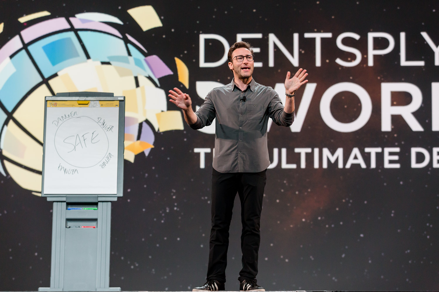 A keynote speaker mid-talk on a wide conference stage, a flip chart beside him and the event branding behind.