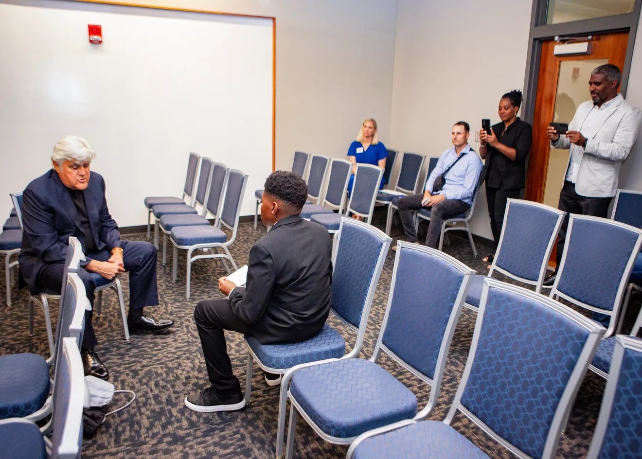 A comedian sits facing a young guest in a meeting room as people nearby watch and film.