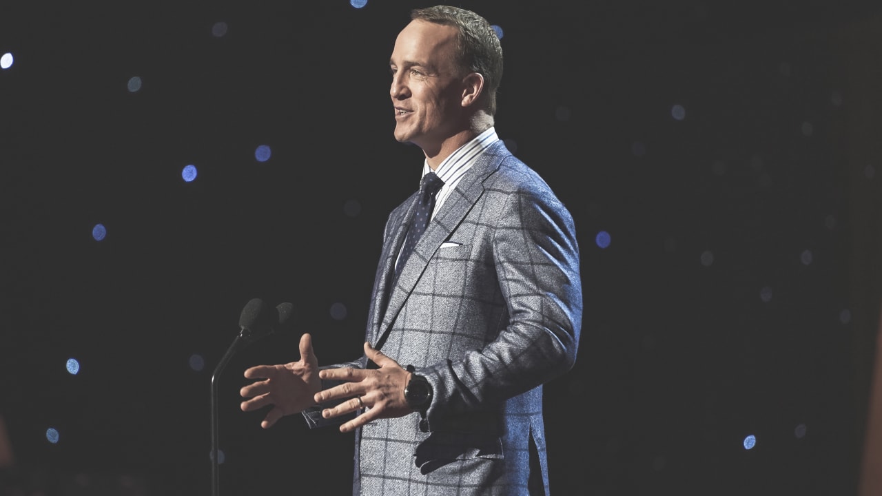 A keynote speaker on a dark stage, mid-sentence, hands open, lights scattered behind him.