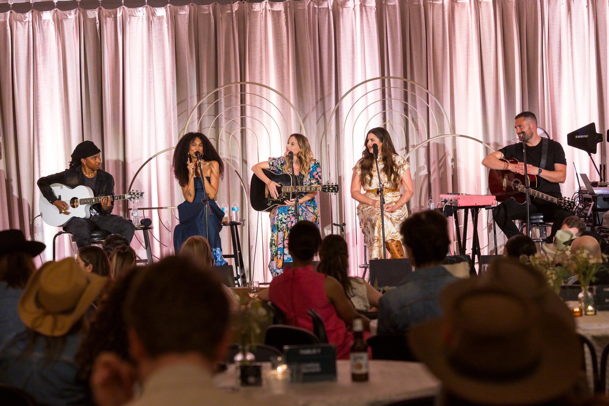 A songwriters' round: five performers seated on stage in front of a seated audience.